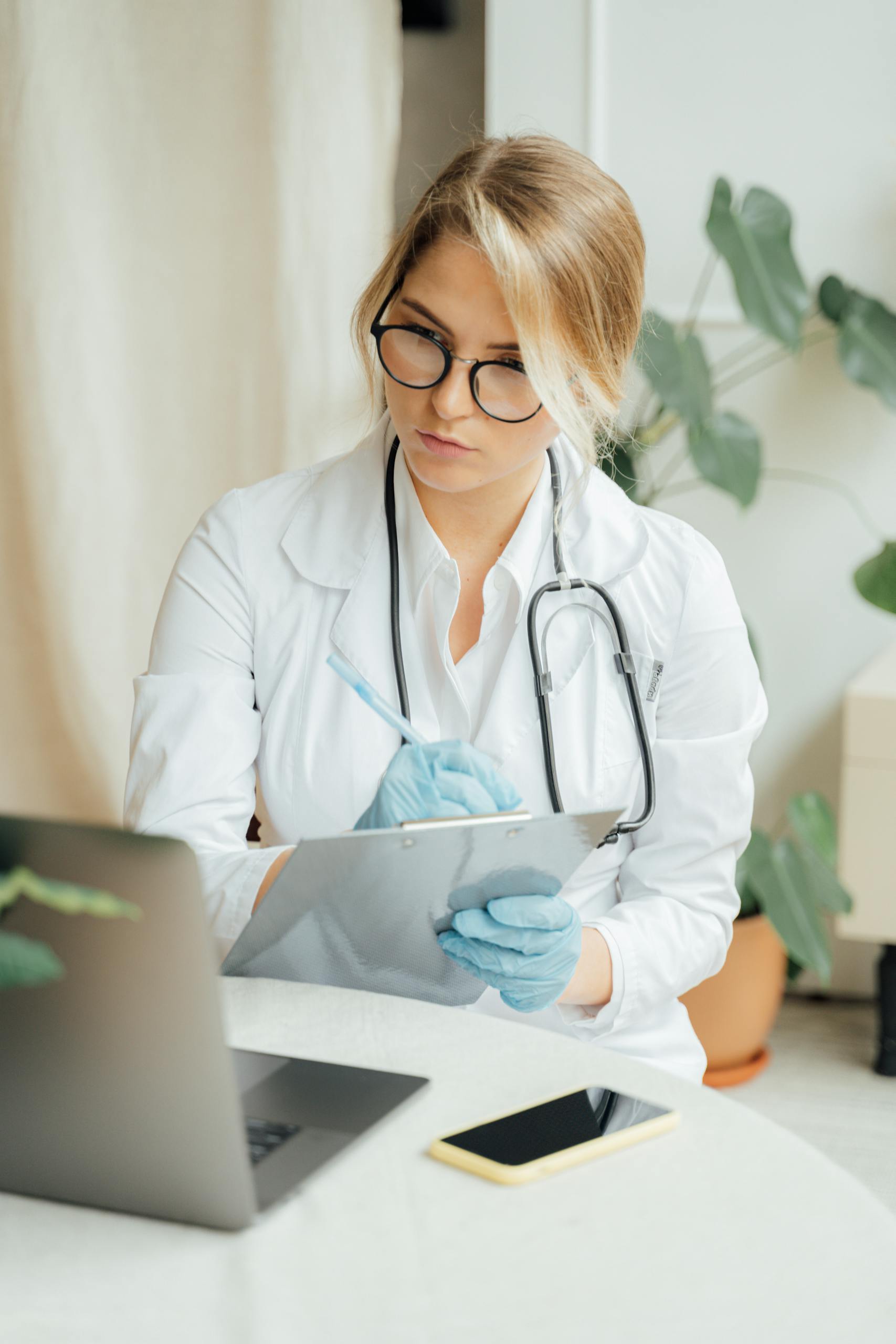 Caucasian female doctor in office attire taking notes on a clipboard with laptop and smartphone on desk.